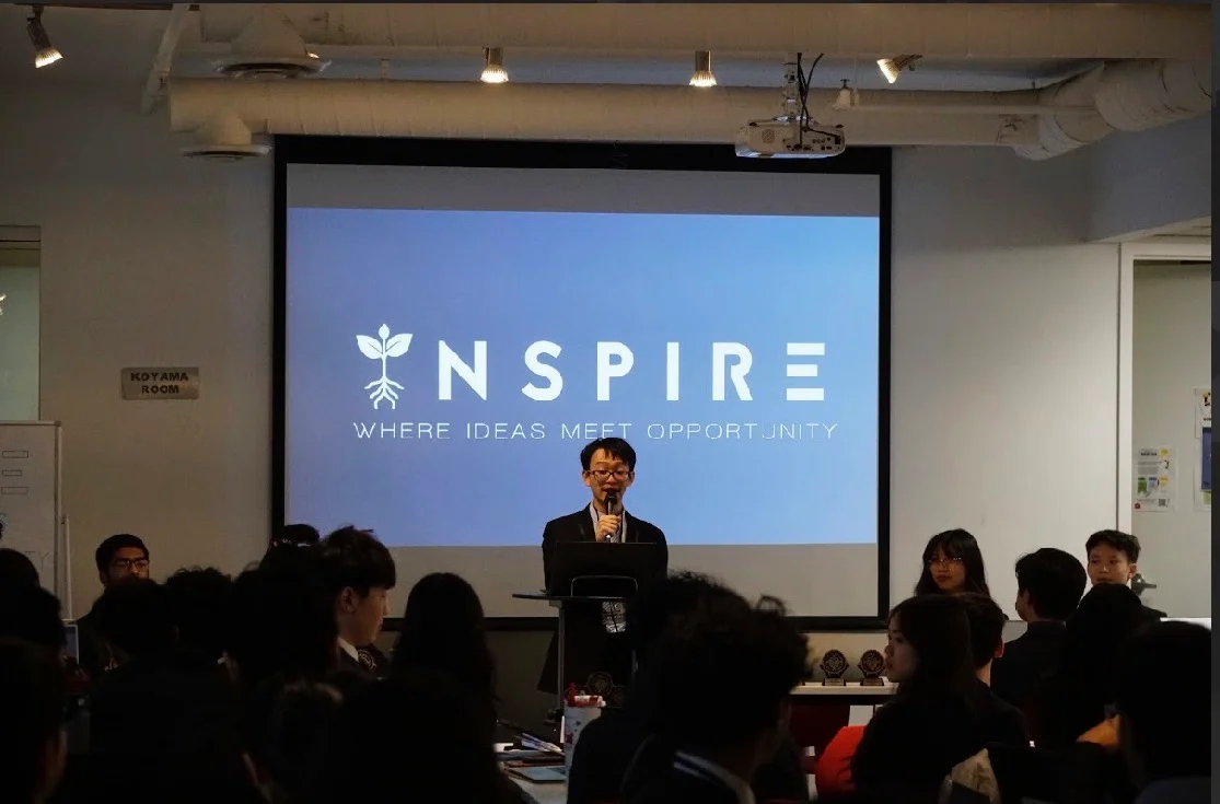 A member of the student team giving the opening address, with the Inspire logo on the screen